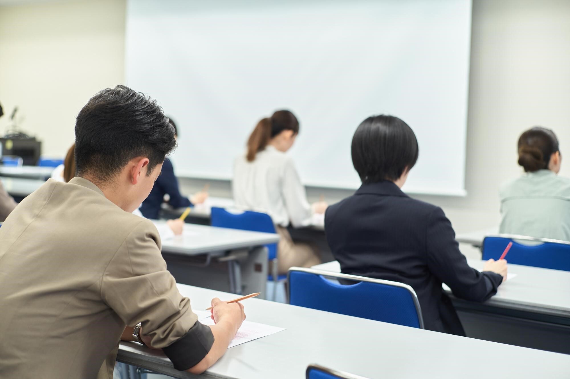 会場で机に向かい試験を受ける多数の男女の後ろ姿