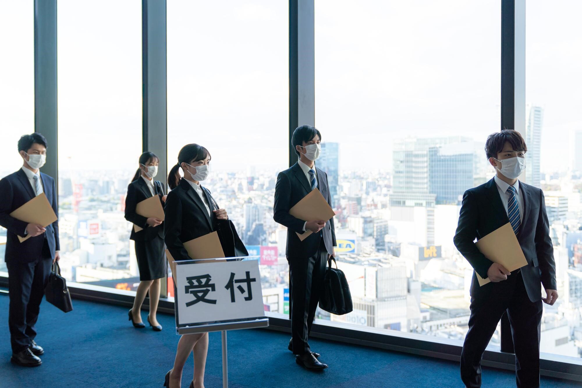 受付の案内看板と手に茶封筒を持ったスーツ姿の複数の男女_