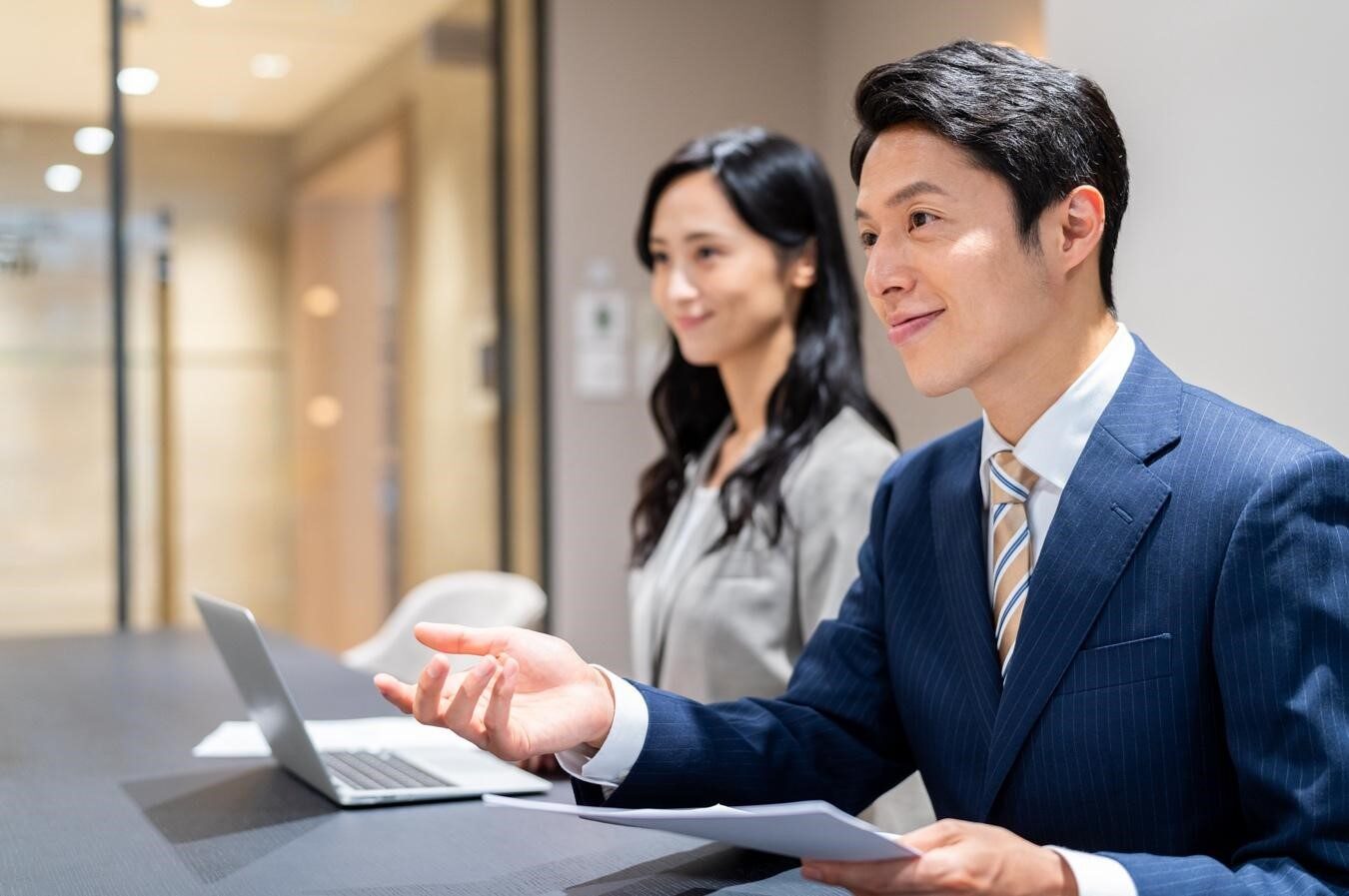 書類を手に笑顔で問いかけるスーツ姿の男性と隣に座る女性