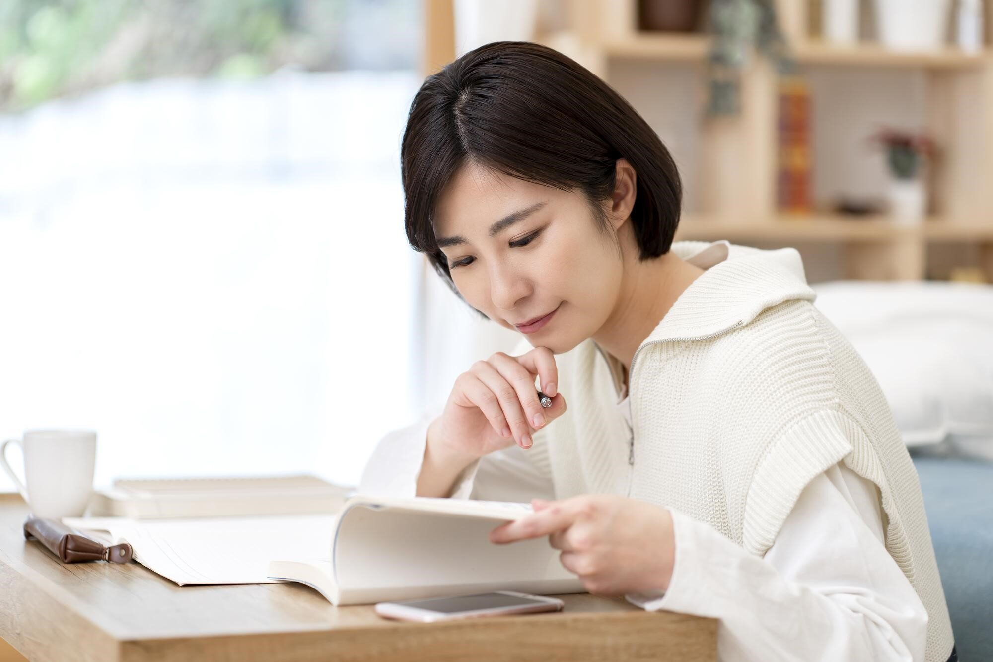 デスクに向かい、ペンを片手に本を開いている女性