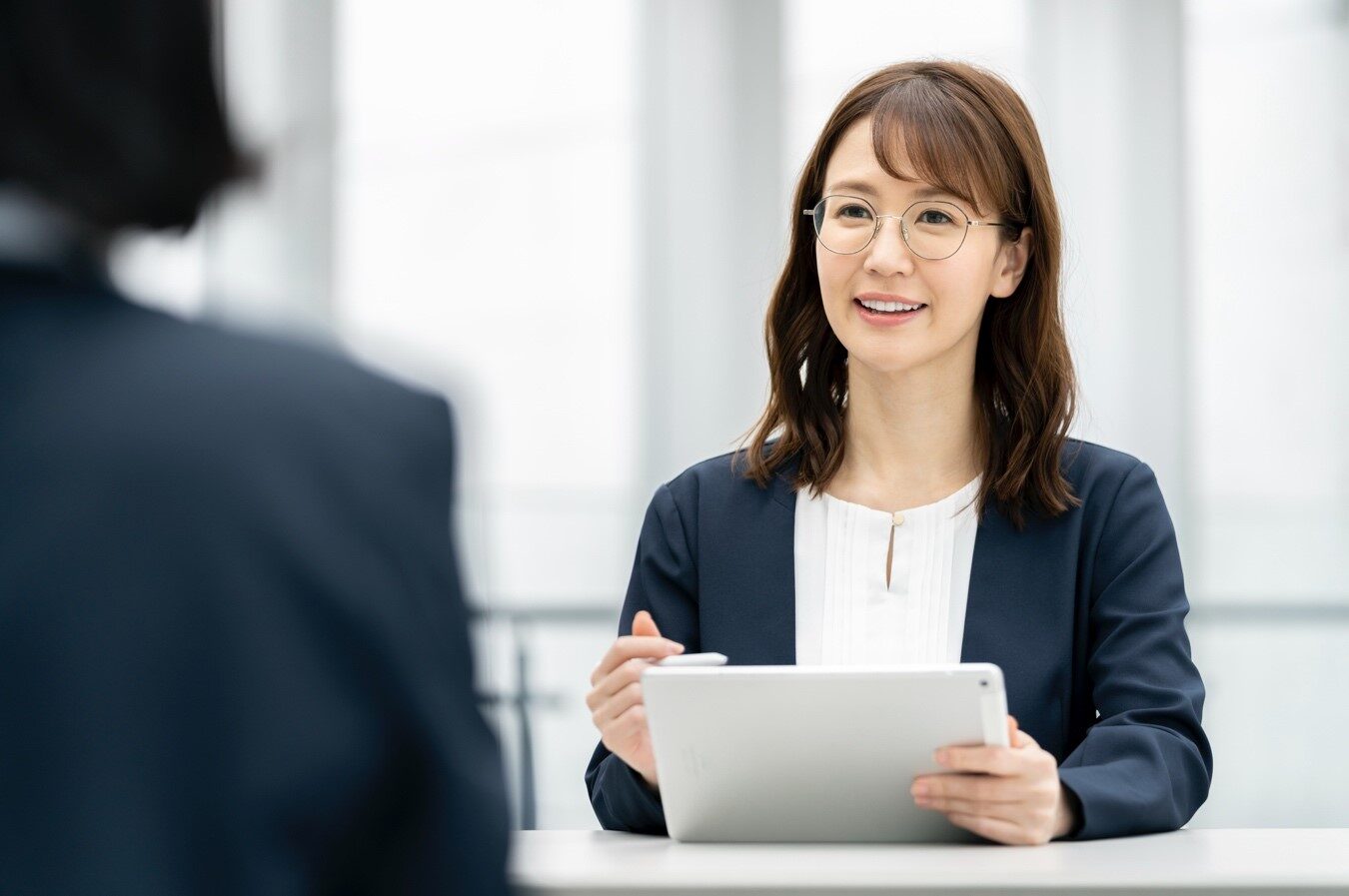 書類とペンを持って向かいに座る人物に話しかける女性