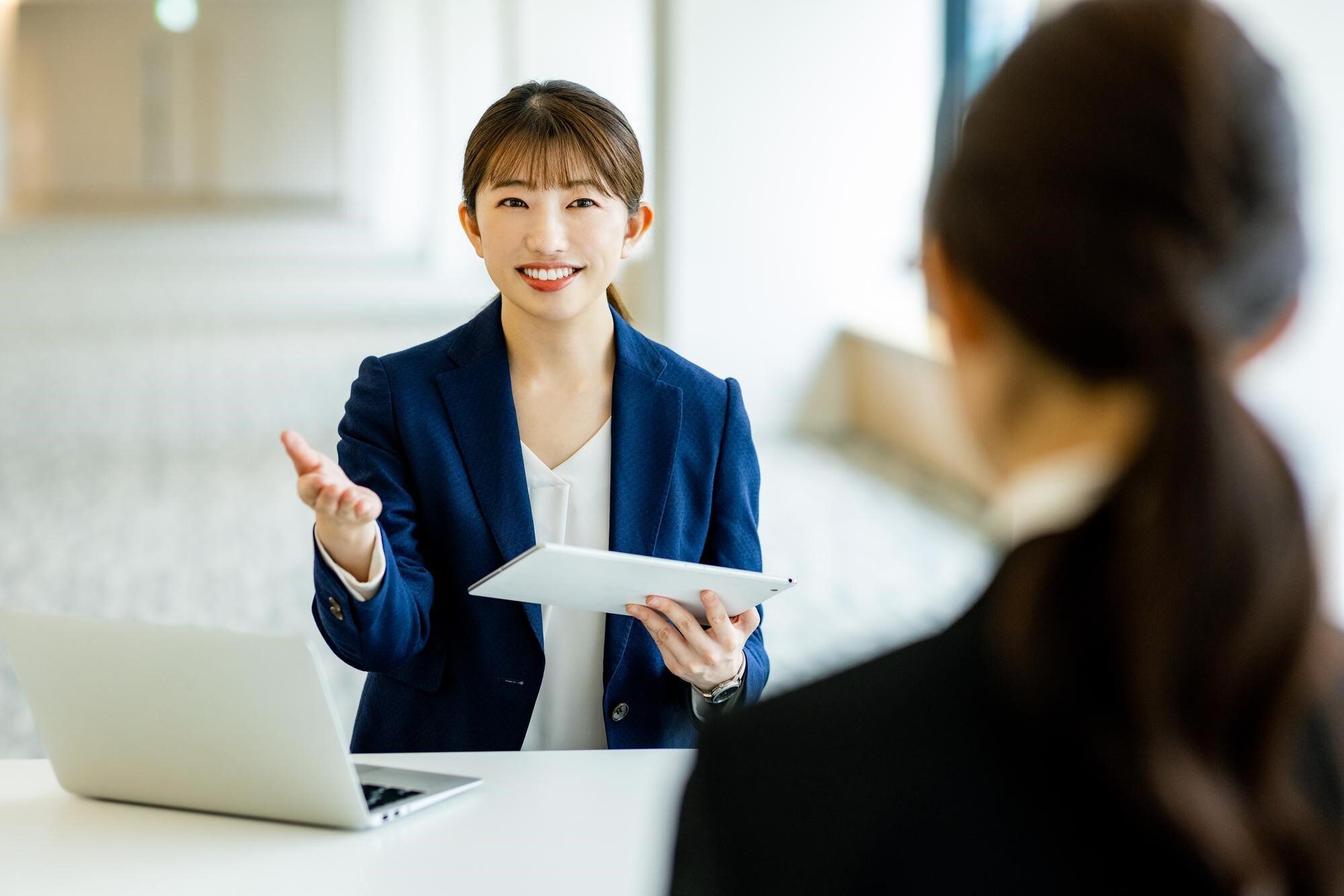 タブレットを手に話す女性と正面に座る人物の後ろ姿